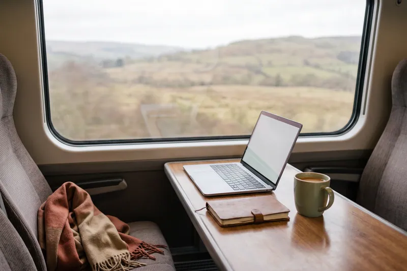 Laptop, Notizbuch und Kaffee auf einem Zugtisch am Fenster.