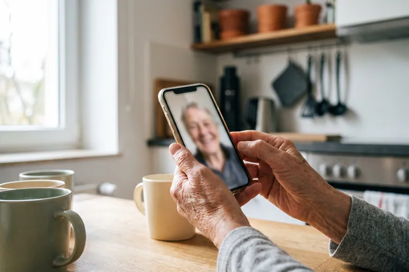 Ältere Hände halten ein Smartphone während eines Videoanrufs in der Küche.