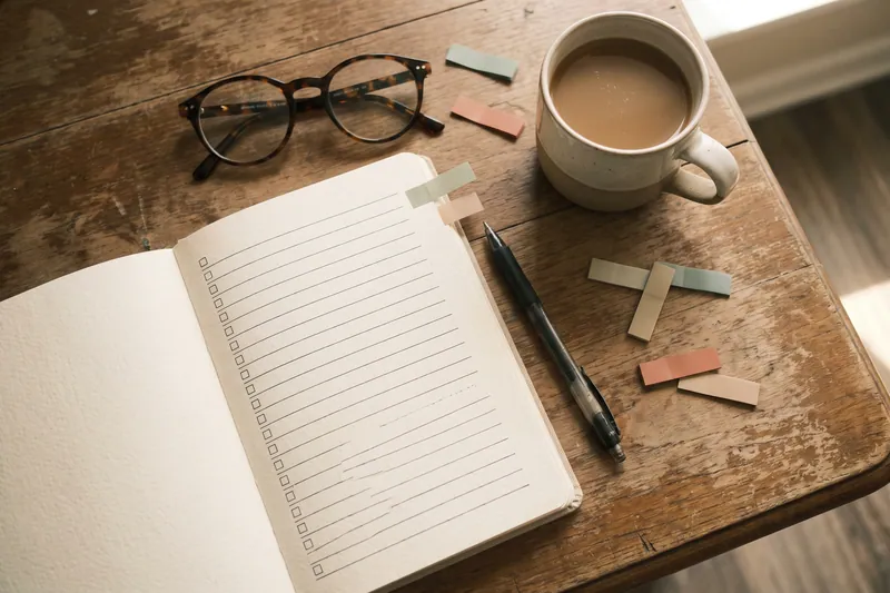 Aufgeklapptes Notizbuch mit leeren Checkboxen, Stift, Lesebrille und Kaffeetasse von oben.